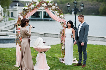 Fototapeta na wymiar Young fashionable lovely couple with speech master of wedding ceremony against decor arch on wedding anniversary and marriage proposal.