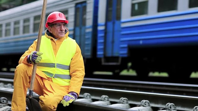 Railway man sits on the rails and looks at the train. Workman with spade on railway track. Railway construction, worker in yellow uniform with shovel in hand sits on railway line