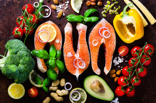 Uncooked salmon fish fillet with aromatic herbs, onion, avocado, broccoli, pepper bell, vegetables on wooden background, top view. Ingredients for cooking on background.