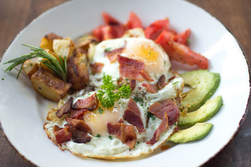 Eggs over medium with a side of avocado, bacon, potato, and tomato 