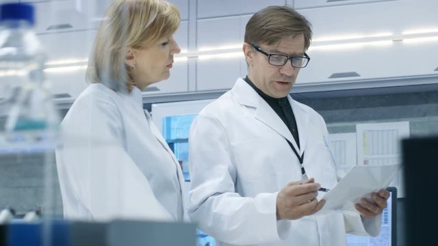 Female and Male Chief Scientists Walking Through Modern Laboratory and Discussing Future Possibilities. Young Woman Assistant Works on Computer at Her Desk. Shot on RED EPIC-W 8K Helium Cinema Camera.