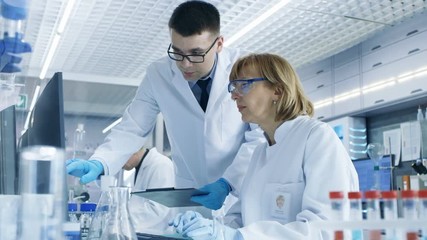 In Modern Laboratory Senior Female Scientist Has Discussion with Young Male Laboratory Assistant. He Shows Her Data Charts on a Clipboard. RED Cinema Camera. 
