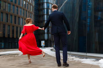 Couple holding hands and dancing in the square in front of the business center. The girl in the red dress and man in elegant black suit.
