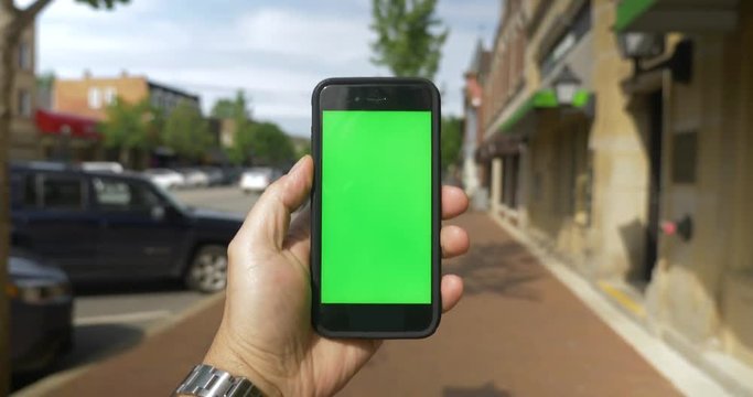 A person's perspective walking in the business district in a small town while holding a green screen smartphone. Optional corner markers included.  	