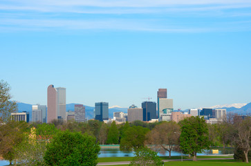 Downtown Denver skyline buildings, on a bright clear summer morning with lake and trees in foreground and snowcapped mountains in background.