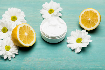 Close-up view of organic cream in container, sliced lemon and chamomiles on blue wooden table