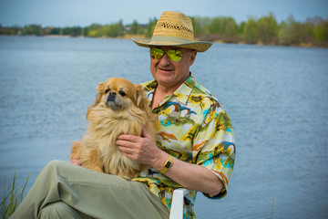 A man of mature age, American appearance, with little red dog resting by blue lake in a Hawaiian shirt, straw hat and green glasses, a cheerful positive and emotional person
