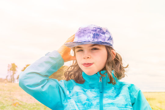 Sunny portrait of cute young girl in blue jacket and cap with wind in her hair, outdoors in open landscape.