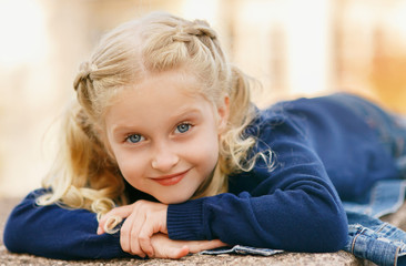 Portrait of a happy liitle girl close-up lying and looking at the camera. Girl with blond hair smiling playfully. Child in a blue sweater and jeans. Autumn toning. Shooting outdoors.