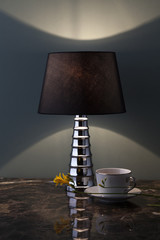 composizione con elegante lampada da tavolo accesa su piano in marmo, tazza da tè e fiore giallo.