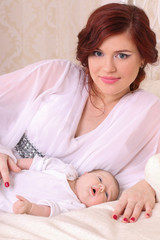Fototapeta na wymiar A young mother in a silk white dress lying on the bed next to her baby and smiling from happiness
