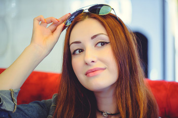 Fototapeta premium young redhead woman in a cafe jabs at his glasses on a red sofa, a playful look