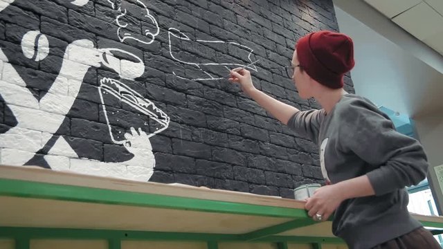 Young female painter making white design on the black brick wall in coffee shop