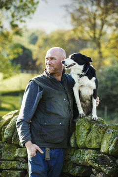 A sheep farmer, shepherd and his sheepdog standing beside him on a dry-stone wall.