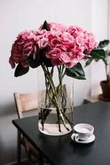 Huge, beautiful and fluffy pink hydrangea on the black wooden coffee table with a coffee cup (filled with pink ranunculus instead of coffee), with a wooden chair on the background.