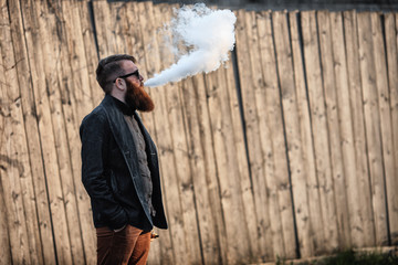 Vape man. Outdoor portrait of a young brutal white guy with large beard and in sunglasses letting puffs out of steam from an electronic cigarette opposite the old wooden fence in the village. Vaping.