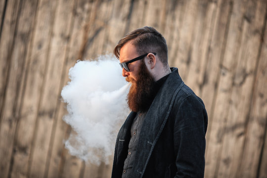 Vape man. Outdoor portrait of a young brutal white guy with large beard and in sunglasses letting puffs out of steam from an electronic cigarette opposite the old wooden fence in the village. Vaping.