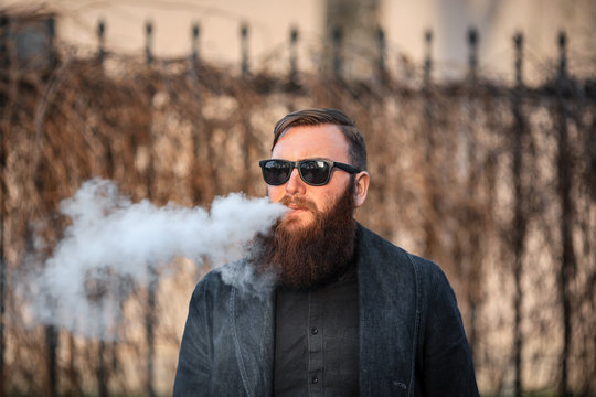 Vape man. Outdoor portrait of a young brutal white guy with large beard and in sunglasses letting puffs out of steam from an electronic cigarette opposite the old wooden fence in the village. Vaping.