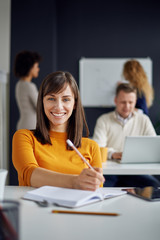 Fototapeta premium Young happy people discussing business standing at the whiteboard in modern light office, beautiful woman sitting at the table at forefront