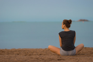 Fototapeta na wymiar Young beautiful woman meditating on the beach in the morning at the dawn of the sun in a gray T-shirt and shorts