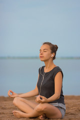 Fototapeta na wymiar Young beautiful woman meditating on the beach in the morning at the dawn of the sun in a gray T-shirt and shorts