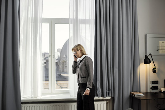 Side view of businesswoman listening to mobile phone standing by window at hotel room
