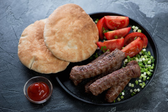 Frying pan with barbecued cevapi or cevapcici sausages, pita bread, tomatoes and onion, studio shot