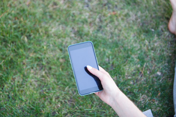 The phone in the girl's hand against the background of the green grass. The girl is writing a message in the park.