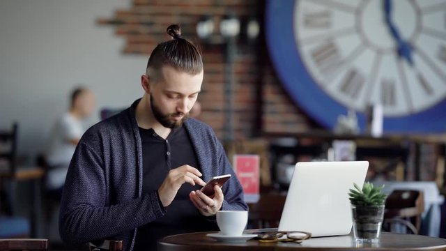An adult guy drinks coffee in a modern cafe, a man with a hair tucked into his tail browses the latest news on the Internet on his mobile phone, there is a laptop on the desk