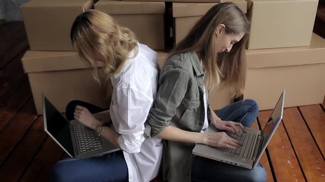 Two Women Are Moving, Sitting Among Cardboard Boxes, Using a Laptop