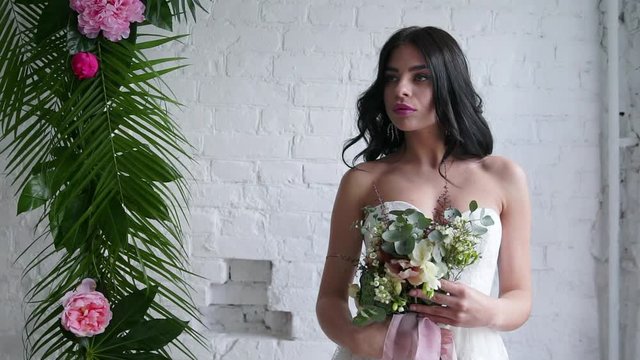 Bride beautiful brunette standing in wedding dress with bouquet near window