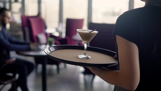 A waiter girl carries a glass of cocktail on a tray for a busy man who spends time in a restaurant at work via the Internet on a laptop