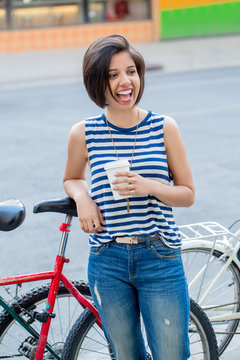 Portrait Of Beautiful Smiling Laughing Young Hipster Latin Colombian Girl Woman With Short Hair Bob In Blue Ripped Jeans, Striped Tshirt, Holding Cup Of Coffee, Leaning  On Bike In City