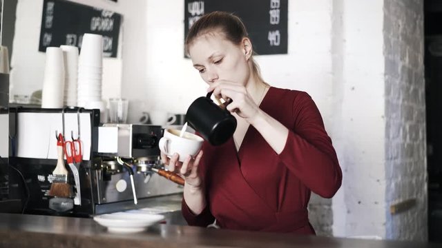 Front view of a woman barista in a red sweater adding milk to a white cup of coffee and putting it on the counter. Locked down real time medium shot