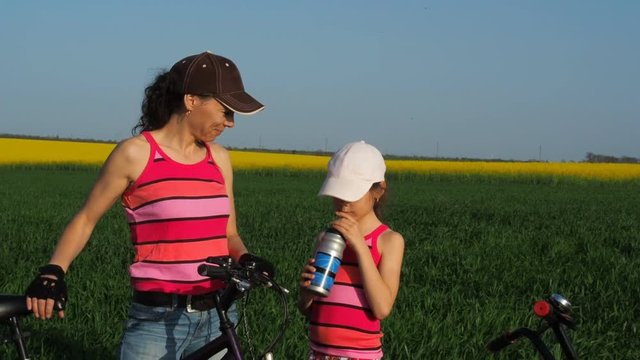 Cyclists drink water. A woman with a child on a bicycle is drinking water. Mom gives her daughter water. The child drinks water. Family in nature.