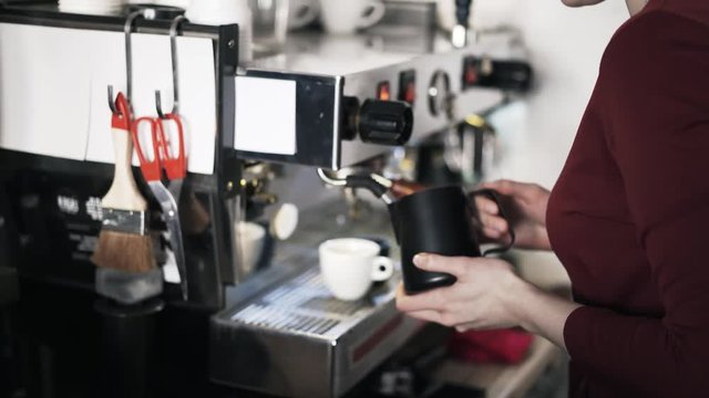 Side view of a woman barista steaming milk for coffee in a black jar. Tilt up real time medium shot