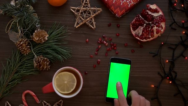 Top view man hands tapping on a smart phone touchscreen with green screen. Christmas decor on the wooden table background. Chroma key. Shot from above