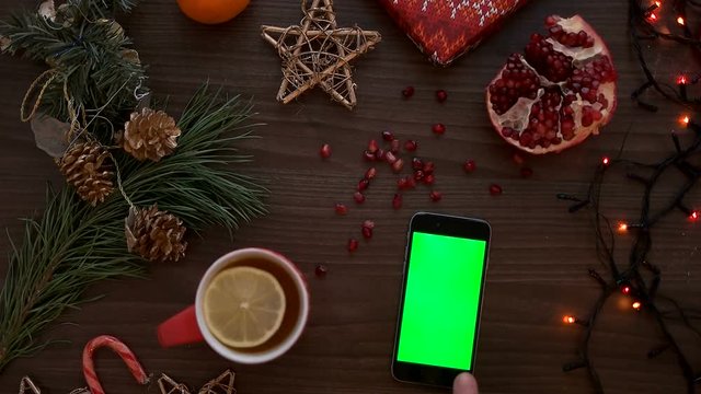 Man finger tapping on a smart phone touchscreen with green screen. Christmas decor on the wooden table background. Chroma key. Top view, Shot from above