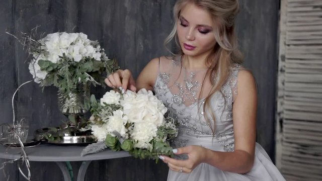 Beautiful blond woman model with a wedding dress sit on a chair with a bouquet of flowers on a gray background