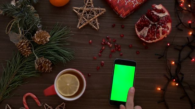 Top view man hand using smart phone with green screen. Finger scrolling pages on touchscreen. Christmas details on wooden table background. Chroma key