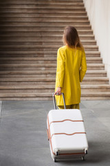 Young woman dressed in yellow suit holding suitcase and standing in front of stairs on railway station. View from the back.