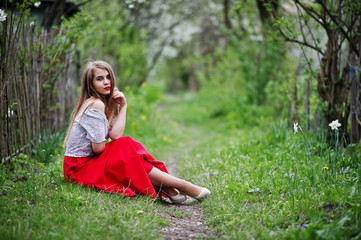 Portrait of sitting beautiful girl with red lips at spring blossom garden on green grass, wear on red dress and white blouse.