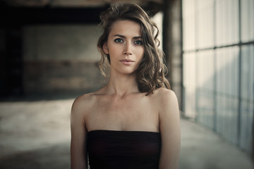 Beautiful girl in in black dress with curly hair posing near the loft window. Woman in retro dress. Worried sensual emotion . Retro fashion