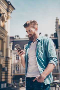 Stylish Young Red Bearded Man In Casual Trendy Jeans Outfit Is Typing A Message On His Phone Outside. He Looks Fashionable And Mature