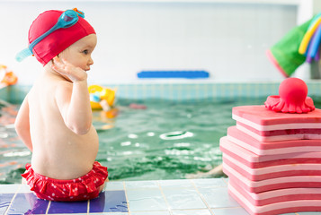 Toddler girl wearing red swimsuit sitting on the edge of a swimming pool near pile of swimming floats looking side
