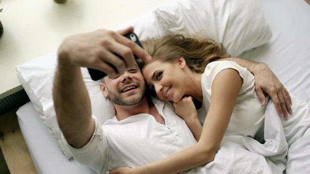Young beautiful and loving couple take selfie picture on smartphone camera and kiss while lying in bed at the morning