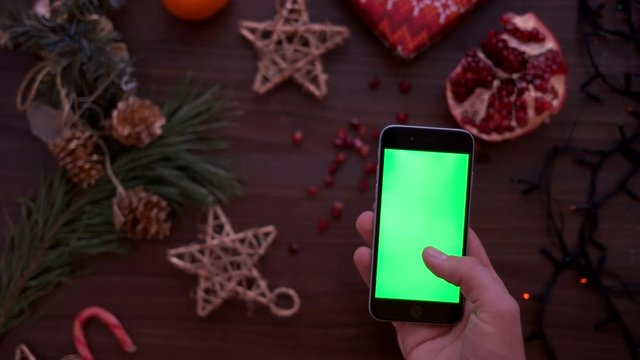 Christmas top view. Man hand using smart phone with green screen. Finger scrolling pages on touchscreen. Christmas details on wooden table background. Chroma key. View from above