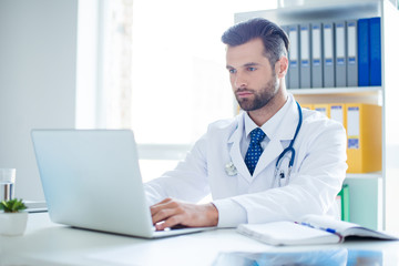 Minded successful doctor sitting at his office and using laptop for his work