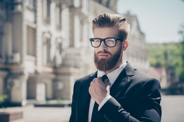 Close up portrait of a successful young red bearded guy in suit and glasses, fixing his tie. So stylish and nerdy. Outdoors on a sunny street