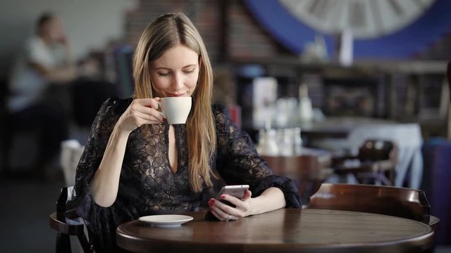 Stylishly dressed woman drinks coffee in a small restaurant, sitting at a wooden table on which there is a cup, the beauty corresponds with her husband on a mobile phone